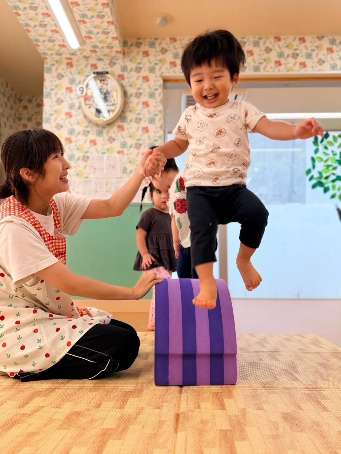 先生と手をつなぎ、紫色の遊具から跳び下りる子ども